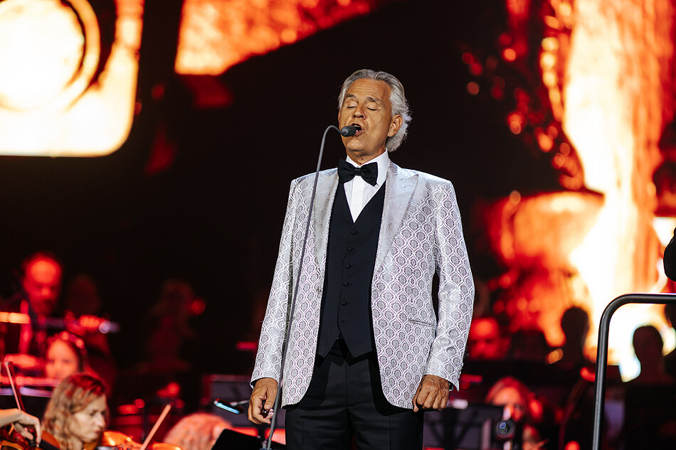 na zdjęciu andrea bocelli w jasnej marynarce, śpiewa do mikrofonu