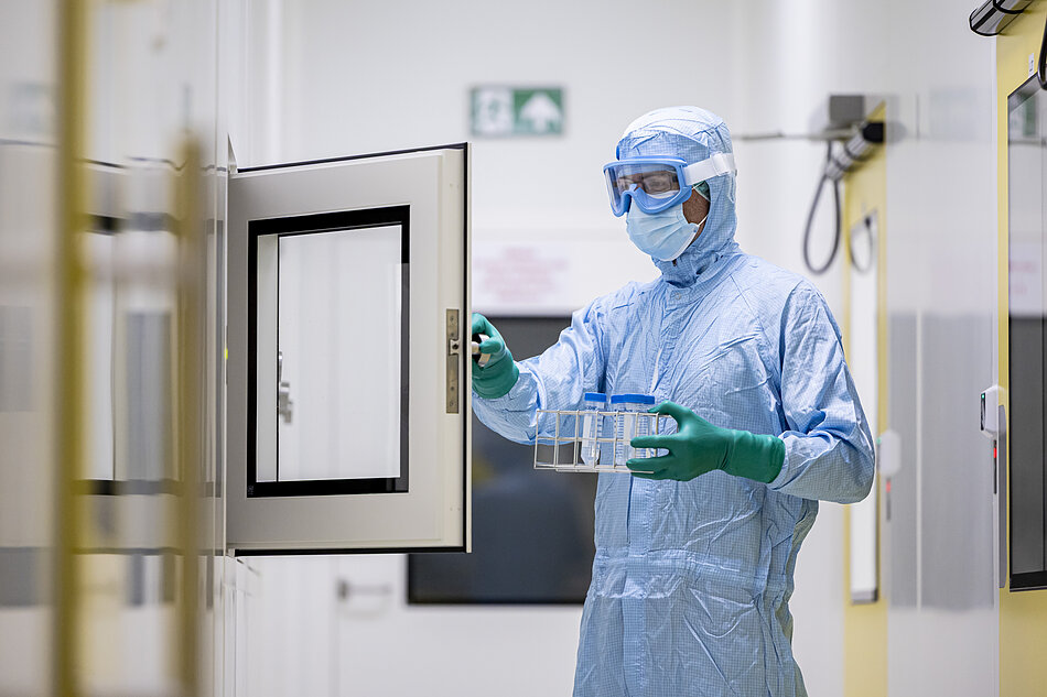  osoba w niebieskim garniturze ochronnym, goglach i rękawiczkach; stoją na korytarzu z białymi ścianami i żółtymi drzwiami; osoba trzyma małą metalową tacykę z kilkoma małymi butelkami i wydaje się, że otwiera drzwi kluczem; na ścianie za osobą znajduje się napis Wyjście awaryjne.