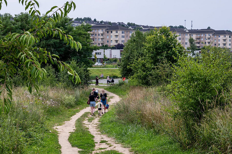 Ludzie idą polna drogą przez zielony teren, w tle widać bloki mieszkalne.