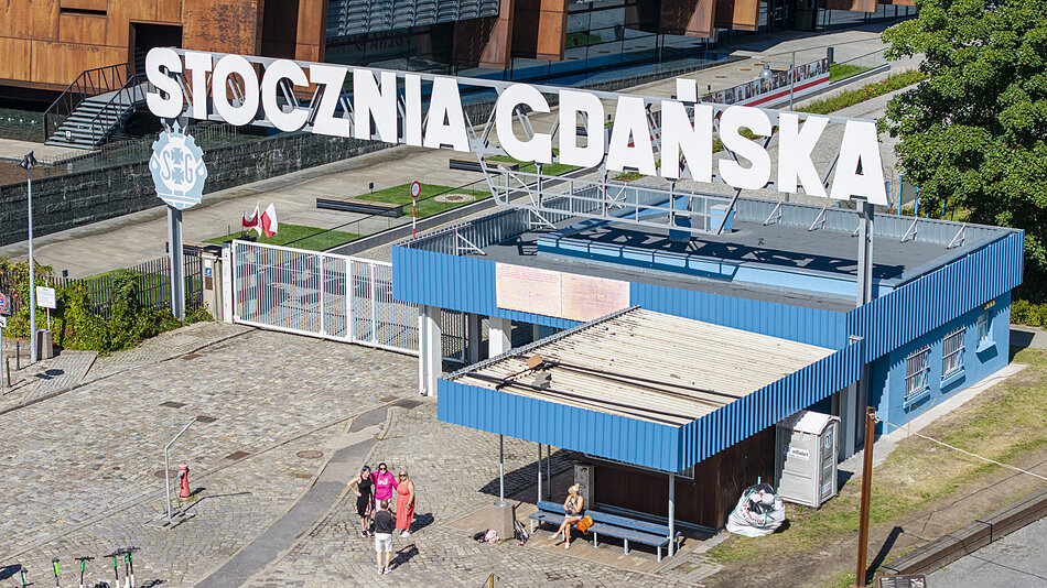  widok z powietrza niebieskiego budynku z napisem Stocznia Cdanskka w białych literach. Budynek wydaje się być dworcem kolejowym lub dworca kolejowego, ponieważ nad wejściem znajduje się duży znak ze słowem STOCZNIA CODANSKA napisanym na nim. Kilka osób chodzi po budynkach, niektóre z nich siedzą na ławkach, a inne na chodniku. Po prawej stronie obrazu widoczne są drzewa i inne budynki.