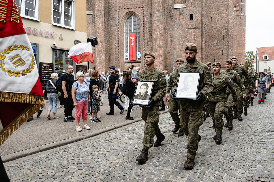 Żołnierze maszerują ulicą Gdańska, Dwóch z przodu trzyma zdjęcia kobiety i mężczyzny w ramkach.