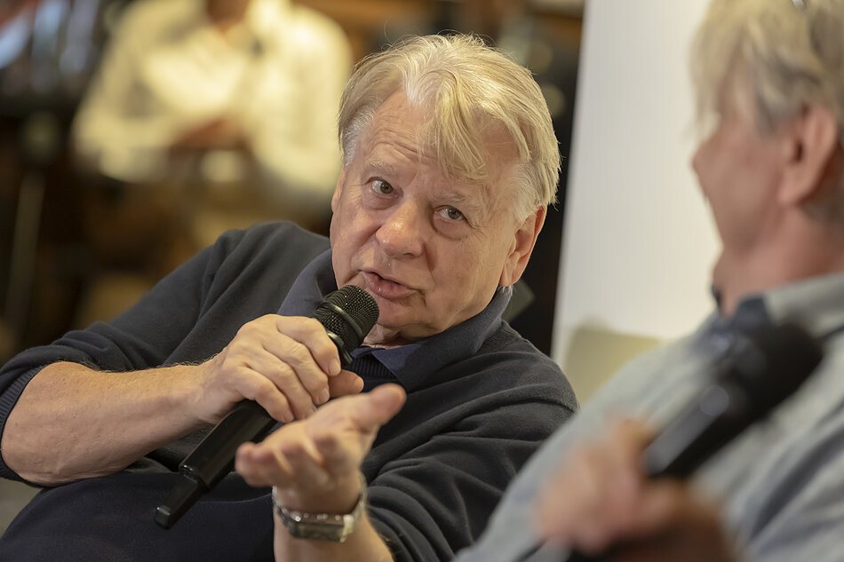 starszy mężczyzna z mikrofonem, siedzi, gestykuluje w kierunku drugiej, rozmytej w kadrze osoby