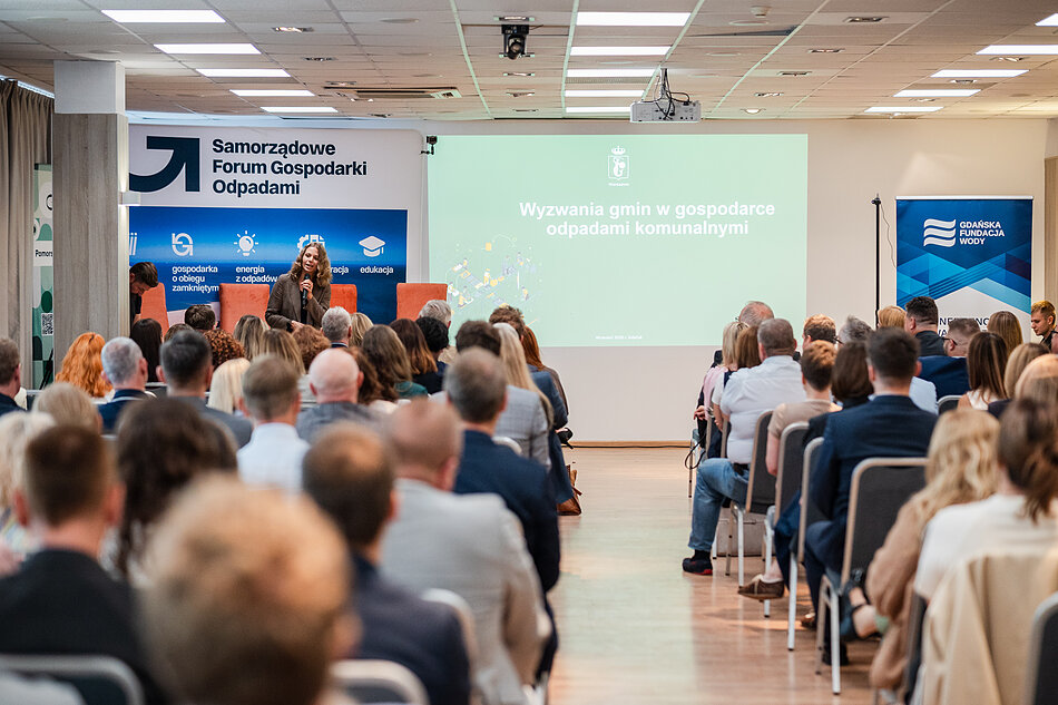 Kobieta przemawia do zebranych osób w sali konferencyjnej.