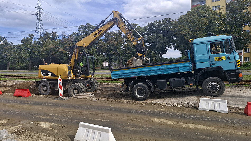 na zdjęciu koparka po lewej i samochód ciężarowy ustawiony do niej tyłem po prawej stronie zdjęcia