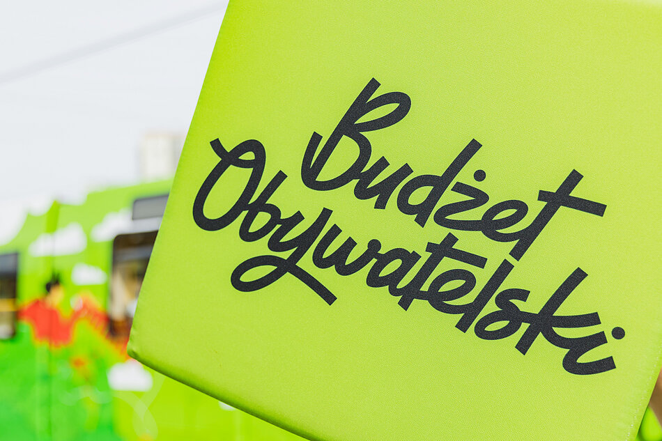 na zdjęciu zielona kostka z czarnym napisem budżet obywatelski, w tle zielony tramwaj