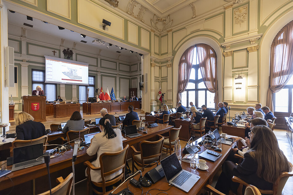 na zdjęciu sala obrad w zabytkowym wnętrzu, widać kilkanaście osób siedzących za stołami tyłem do fotografującego