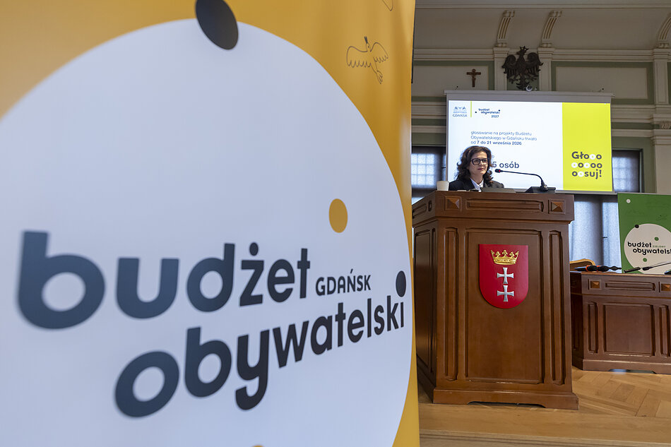 na pierwszym planie z lewej żółta stojąca plansza, w tle po prawej kobieta stojąca za drewnianą mównicą