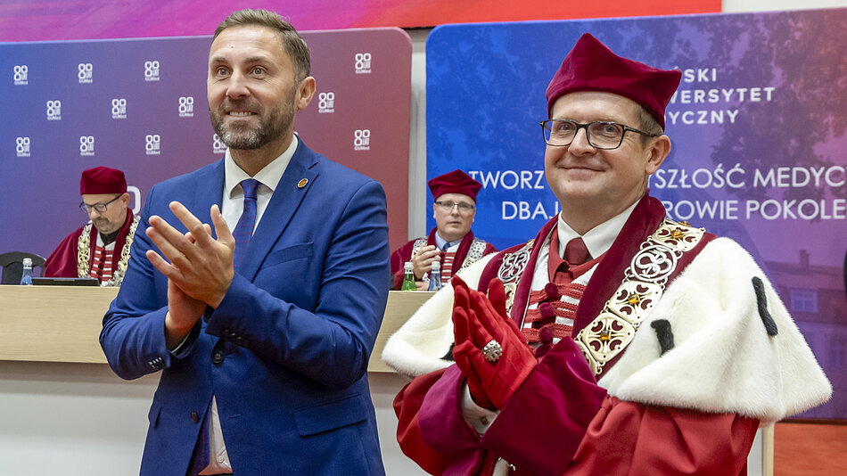  Dwóch mężczyzn stojących przed podium z banerem za sobą, mężczyzna po lewej stronie nosi niebieski garnitur, ma brodę, drugi ubrany w tradycyjny strój uczelniany z togą i biretem