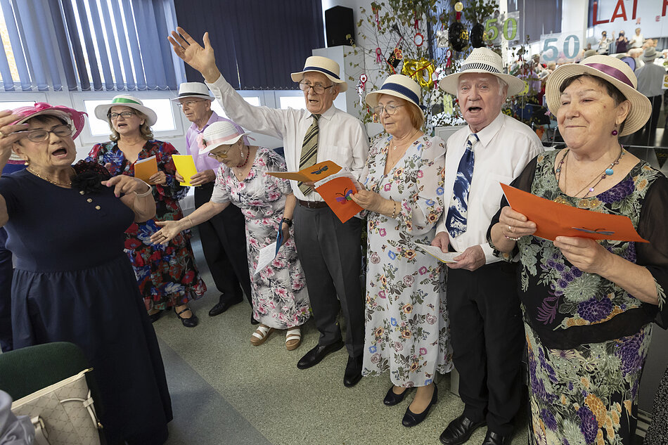 na zdjęciu grupa seniorów i seniorek, stoją, część śpiewa, mężczyzna unosi do góry rękę