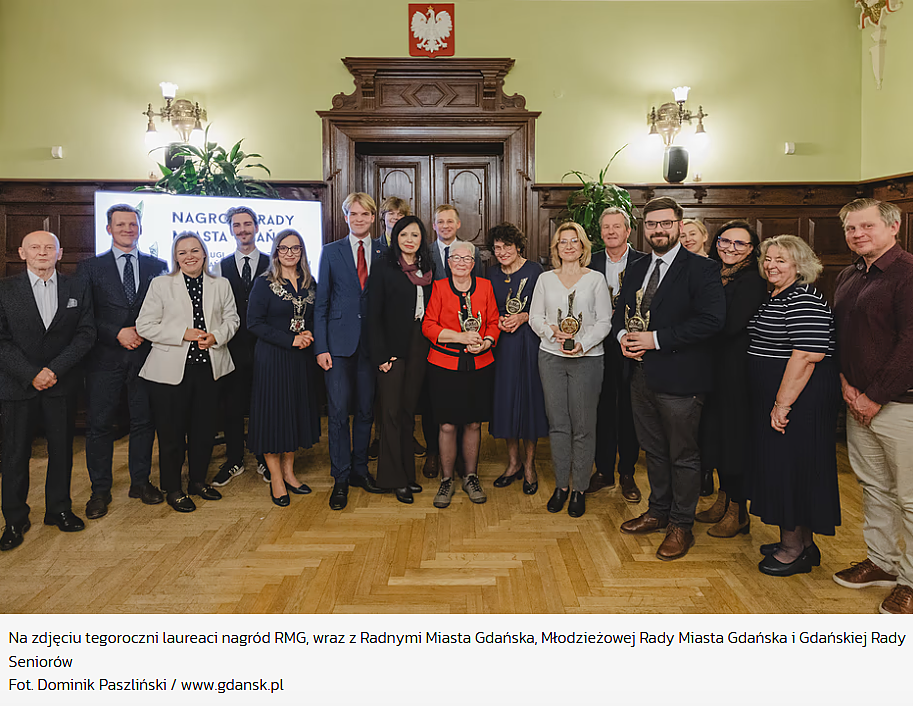 **Alt text:** Zdjęcie grupowe przedstawiające tegorocznych laureatów nagród Rady Miasta Gdańska (RMG) wraz z radnymi Miasta Gdańska, Młodzieżowej Rady Miasta Gdańska oraz Gdańskiej Rady Seniorów. Uczestnicy spotkania stoją w eleganckiej sali z drewnianymi panelami i godłem Polski na ścianie, a niektórzy z nich trzymają w rękach statuetki. Na dole widoczny podpis: „Fot. Dominik Paszliński / www.gdansk.pl”.