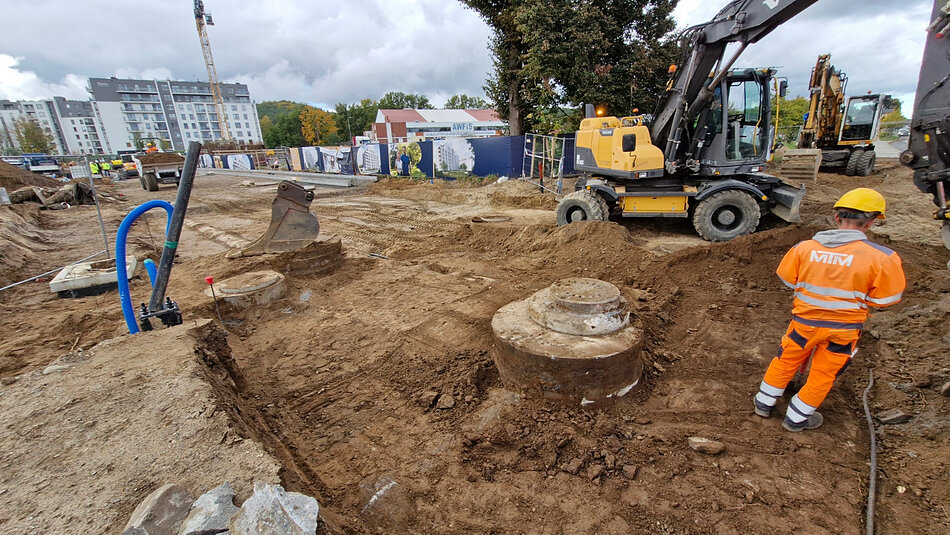 na zdjęciu plac budowy, rozkopana droga gruntowa, widać robotników w kaskach i koparkę