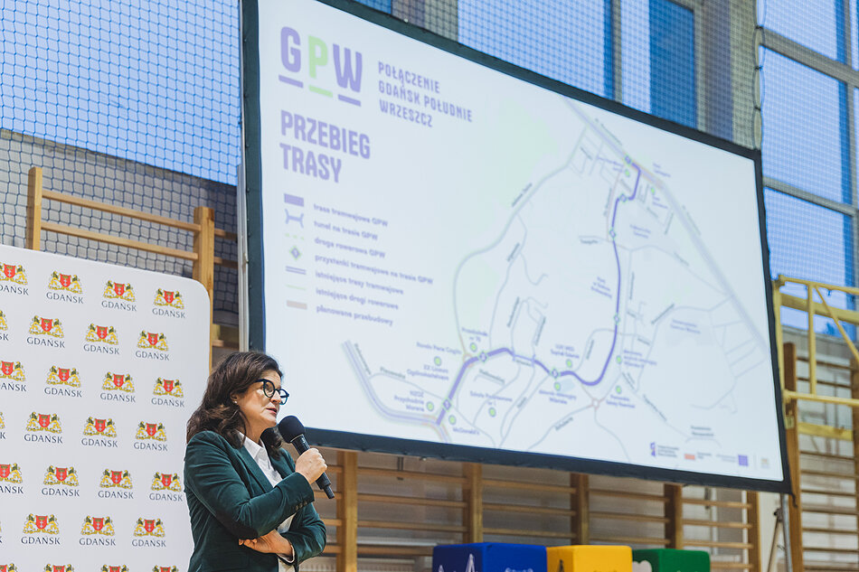 na zdjęciu prezydent dulkiewicz mówi do mikrofonu, za nią wyświetla się prezentacja graficzna