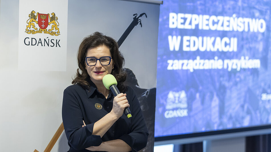 Kobieta z ciemnymi włosami przemawiająca do mikrofonu, za nią plansza z napisem BEZPIECZEŃSTWO W EDUKACJI - zarządzanie ryzykiem