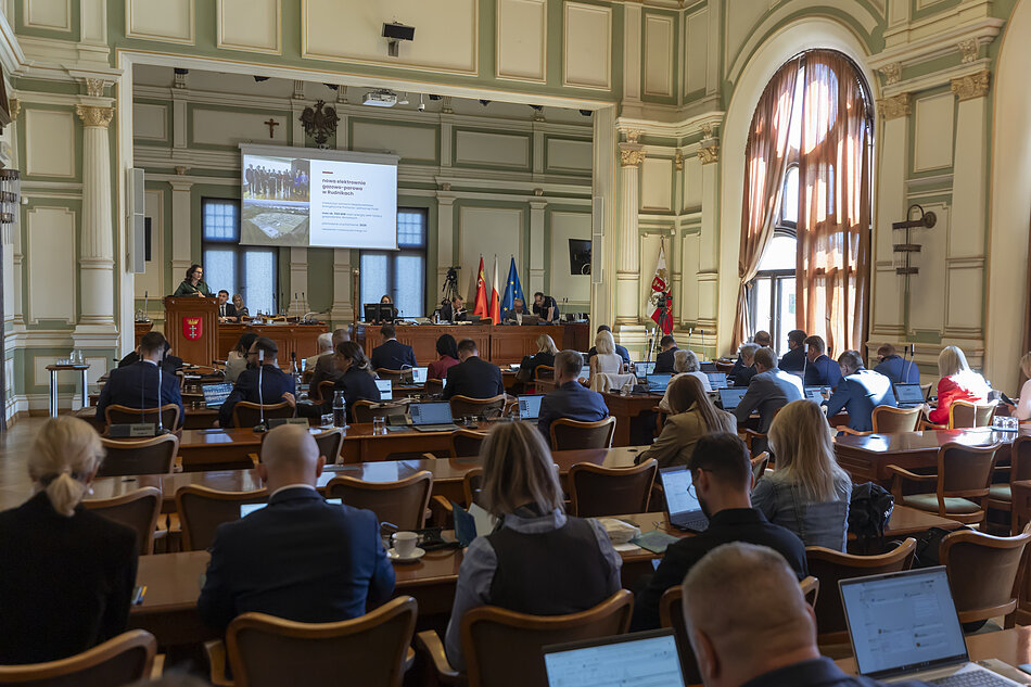 radni siedzą przy stołach, w tle podest z mównicą i ekranem prezentacyjnym