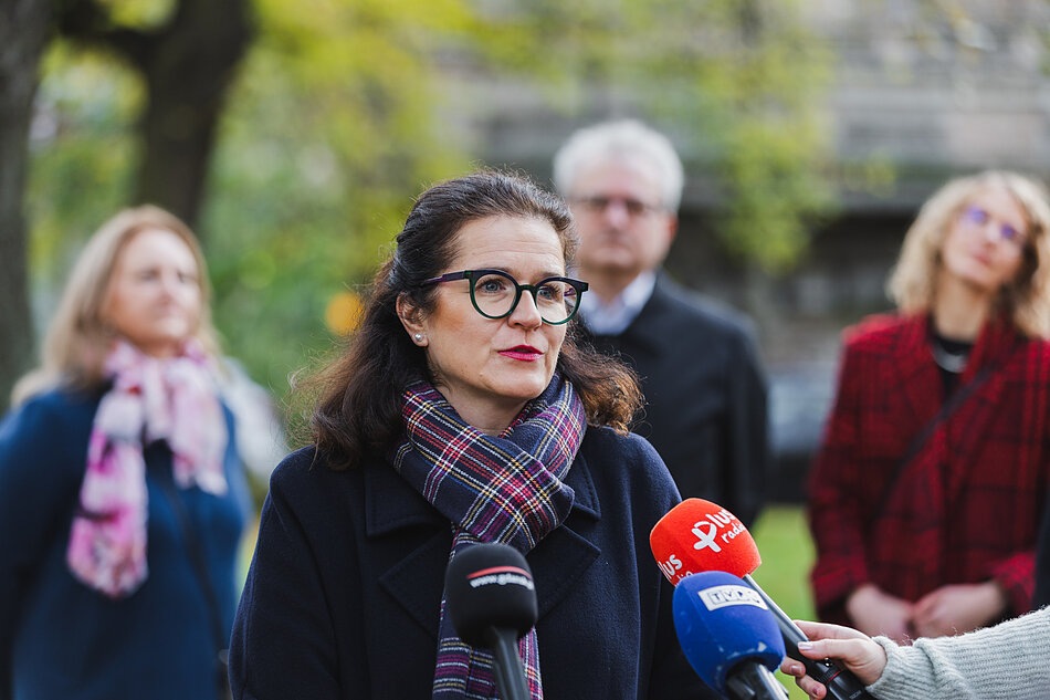 na zdjęciu prezydent gdańska w ciemnym płaszczu mówi do mikrofonów dziennikarskich