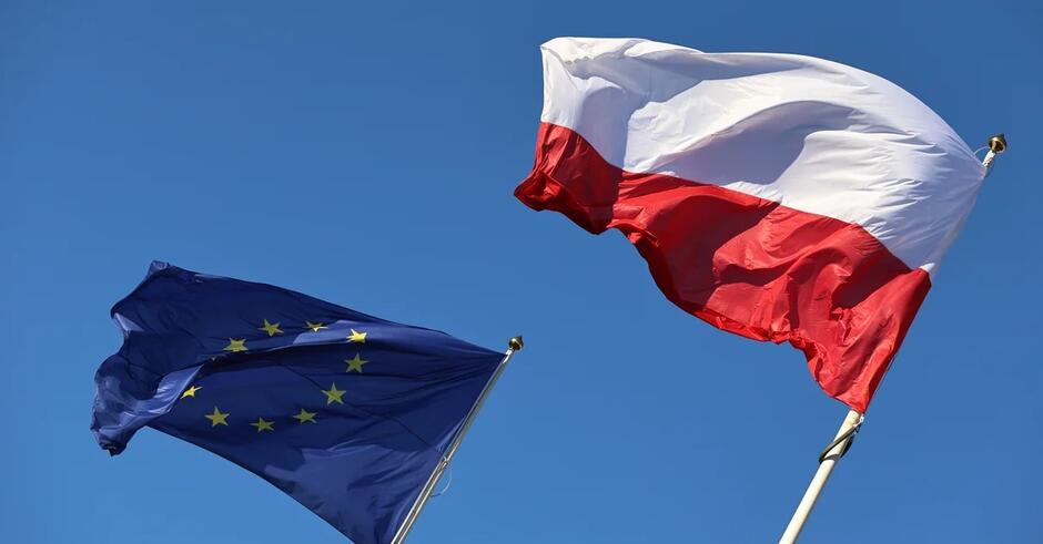 Na zdjęciu widnieją powiewające flagi - Unii Europejskiej oraz Polski - na tle nieba.