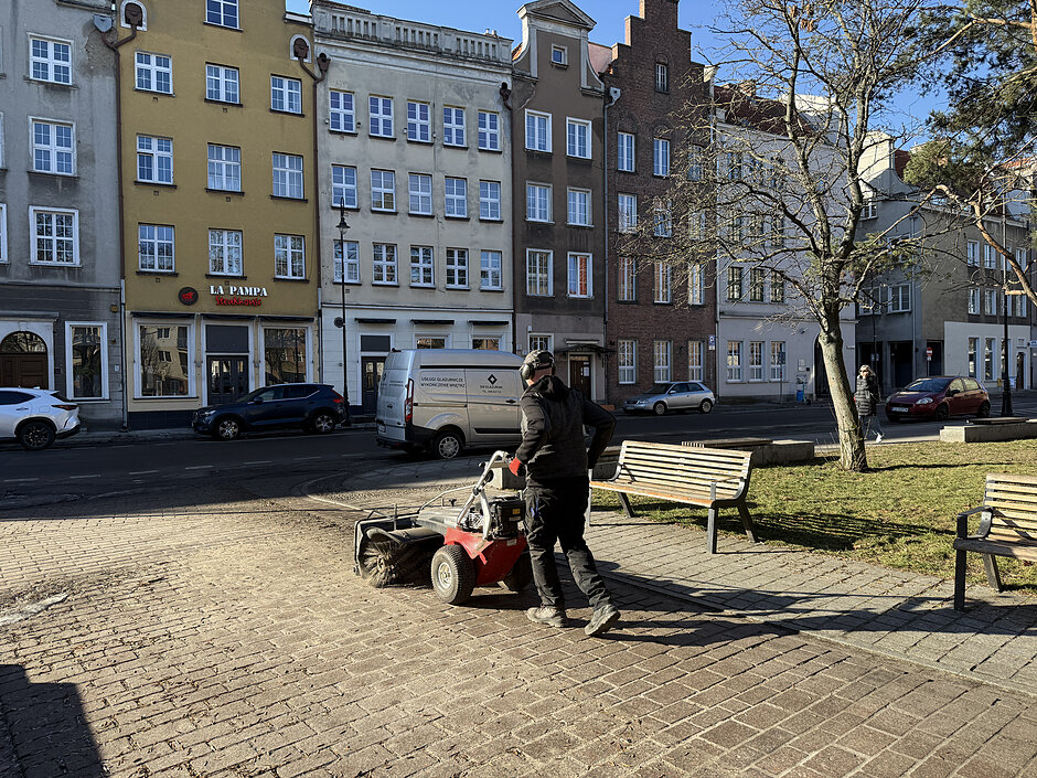 Na zdjęciu widoczny jest pracownik oczyszczający nawierzchnię placu za pomocą specjalnej maszyny