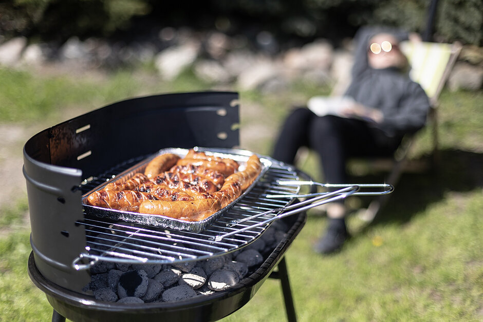 Na zdjęciu widoczny jest grill, na którego ruszcie opiekają się kiełbaski. W tle widać osobę leżącą na leżaku.