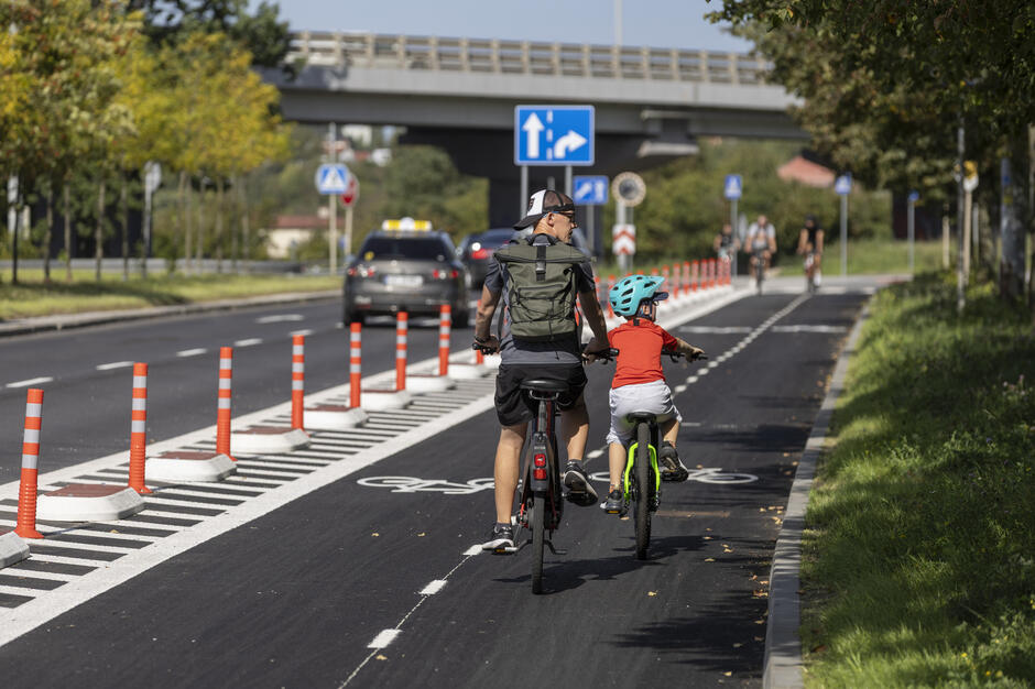 Dwóch rowerzystów jadących po drodze rowerowej wydzielonej z jezdni, w słoneczny dzień. W oddali widać estakadę oraz samochód