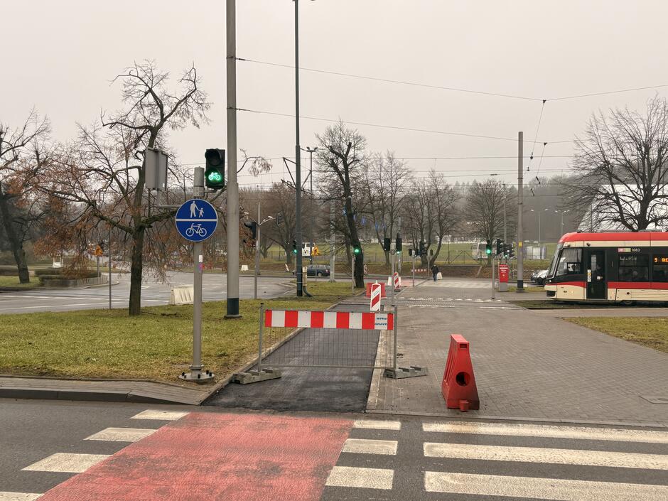 Widok na przejście dla pieszych i przejazd rowerowy zimową porą, w oddali widać tramwaj oraz drzewa bez liści. Na drugim planie Plac Zebrań Ludowych