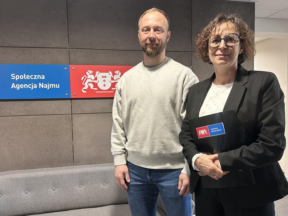 Po lewej stoi mężczyzna w jasnej bluzie i jeansach, po prawej kobieta w ciemnym stroju i okularach, a w tle widać logo Społecznej Agencji Najmu.