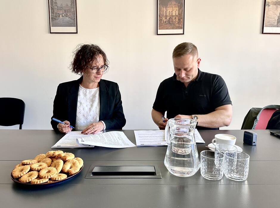 Po lewej siedzi kobieta w ciemnej marynarce, jasnej bluzce i okularach, po prawej mężczyzna w ciemnej koszulce, a przed nimi na stole leżą dokumenty, stoi dzbanek z wodą i widać talerz z ciastkami.