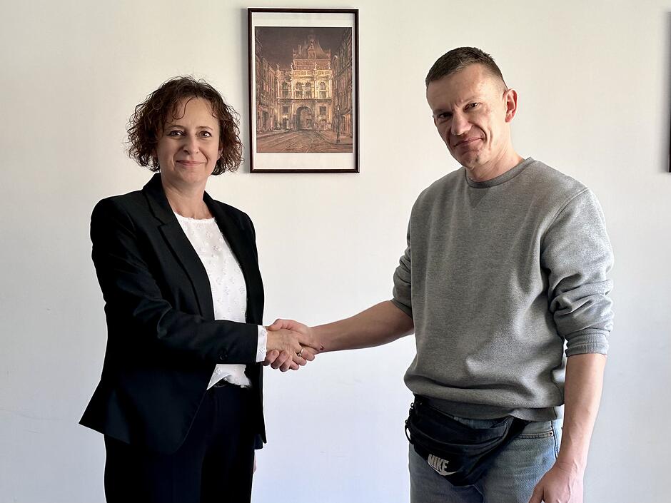 Po lewej stoi kobieta w ciemnej marynarce i jasnej bluzce, po prawej mężczyzna w szarej bluzie i jeansach, a oboje podają sobie ręce.