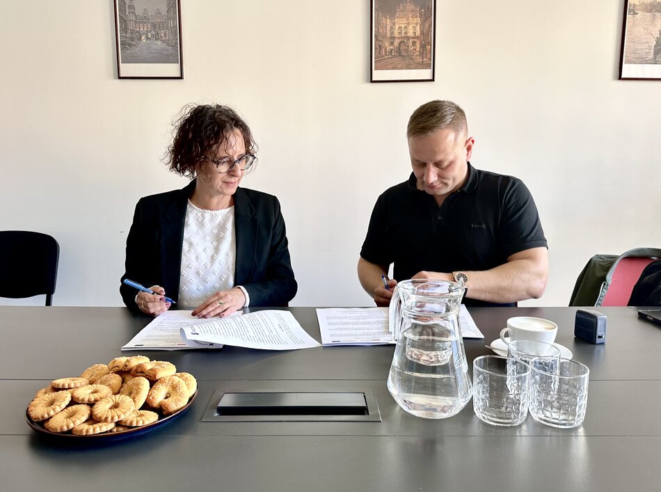 Po lewej siedzi kobieta w ciemnej marynarce, jasnej bluzce i okularach, po prawej mężczyzna w ciemnej koszulce, a przed nimi na stole leżą dokumenty, stoi dzbanek z wodą i widać talerz z ciastkami.