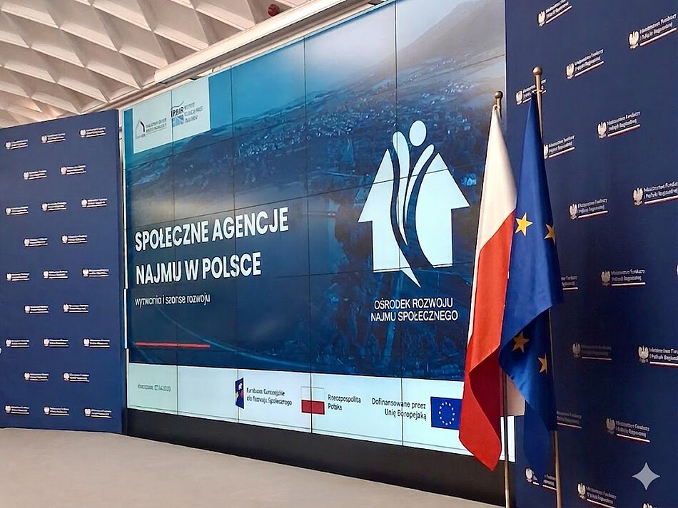 Wyświetlacz pomiędzy dwoma planszami z logo Ministerstwa Funduszy i Polityki Regionalnej. Na pierwszym planie flaga Polski i Unii Europejskiej