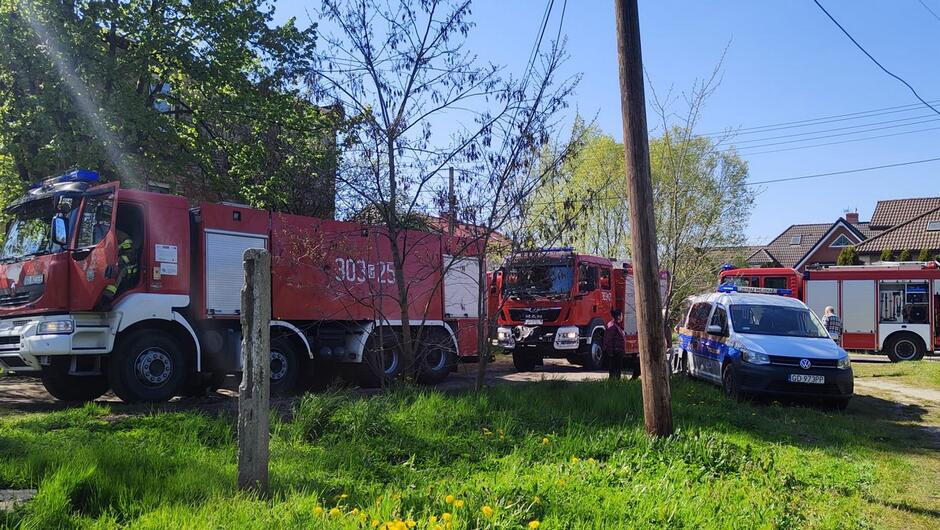 radiowóz straży miejskiej trzy wozy straży pożarnej stoją na trawniku podczas akcji gaszenia pożaru.