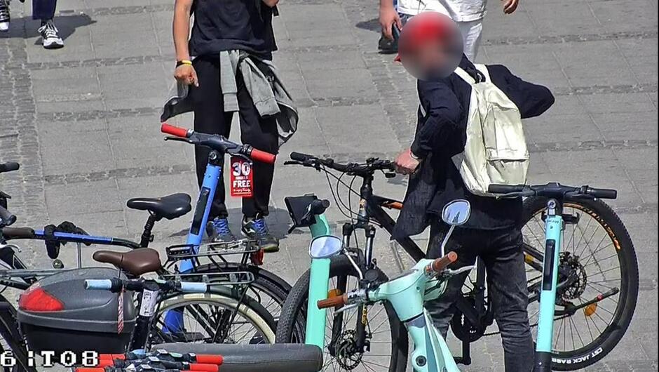 Mężczyzna zabiera zaparkowany rower przy stojaku. W tle inne osoby 