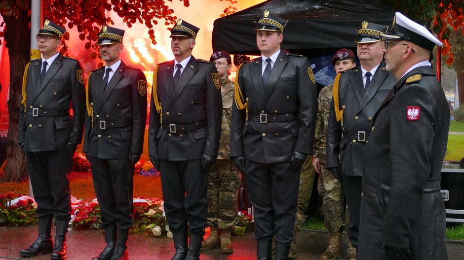 delegacja pięciu gdańskich strażników stoi na baczność, w tle czerwone race