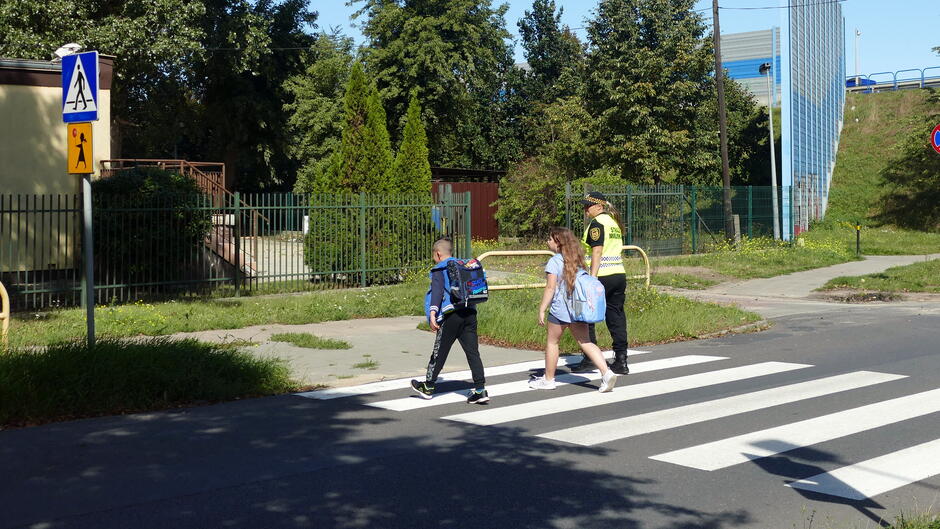 Dwoje dzieci i strażniczka miejska przechodzą przez przejście dla pieszych.