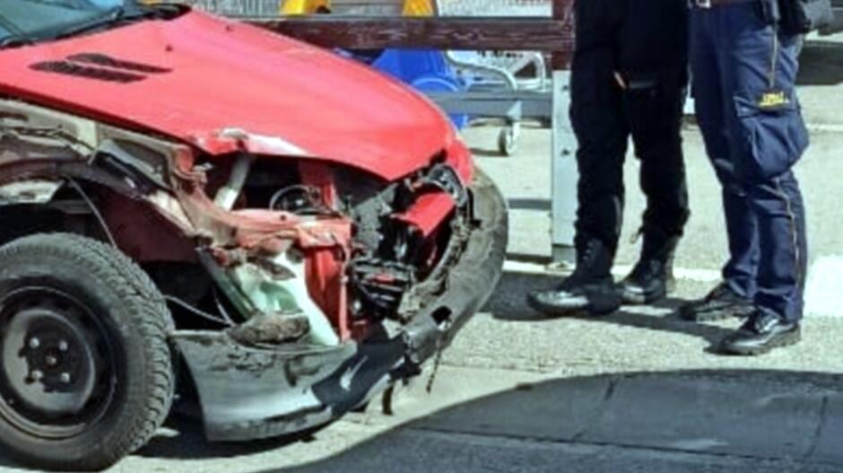 uszkodzony przod auta osobowego, przed nim nogi stojacych straznikow