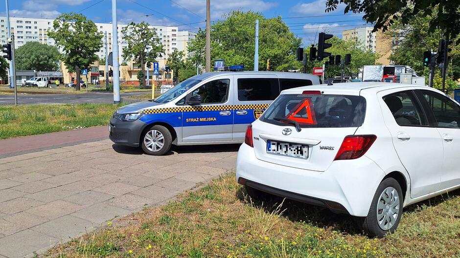 białe auto na poboczu. za tylna szybą trójkąt ostrzegawczy. w tle radiowóz strażników miejskich.