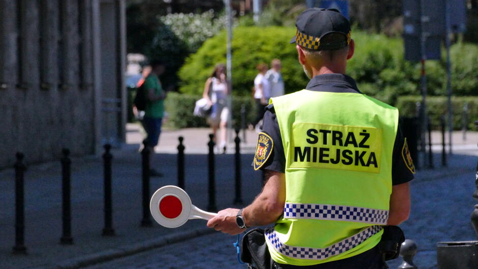 straznik tyłem w kamizelce odblaskowej, w lewej dłoni trzyma lizak