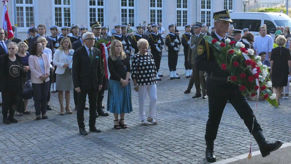 Strażnik miejski a za nim delegacja składają wieniec kwiatów. 