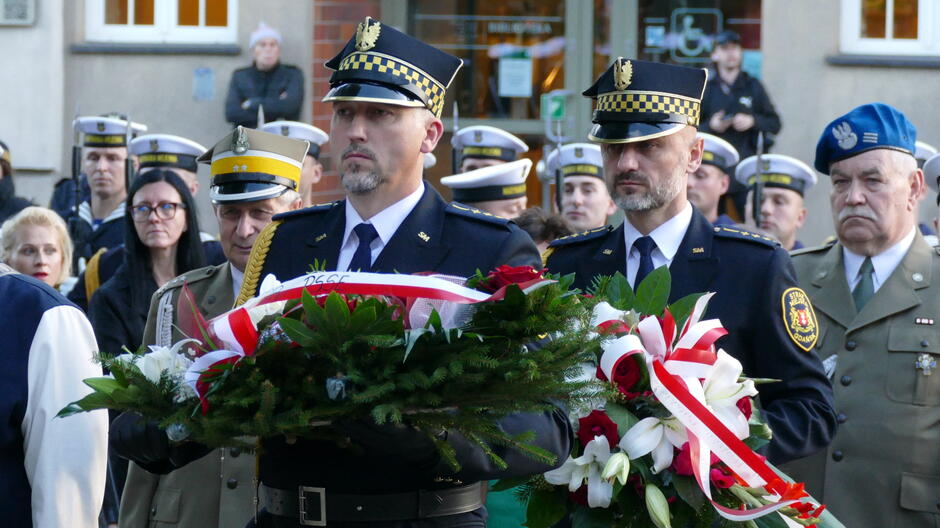 strażnicy trzymają wiązanki kwiatów. za nimi uczestnicy.