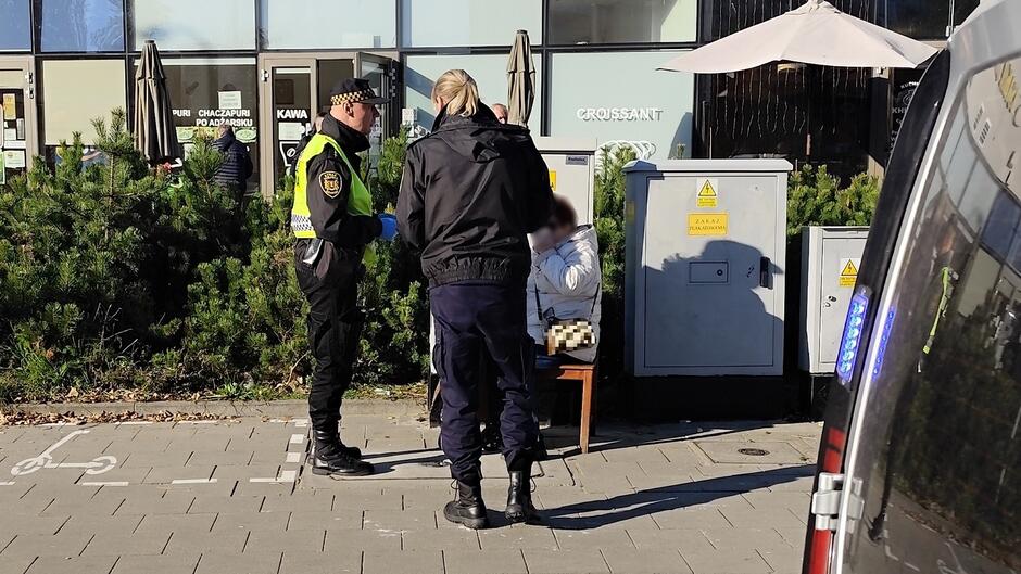 starsza pani z opatrunkiem siedzi na krześle, przy niej stoi strażnik i strażniczka