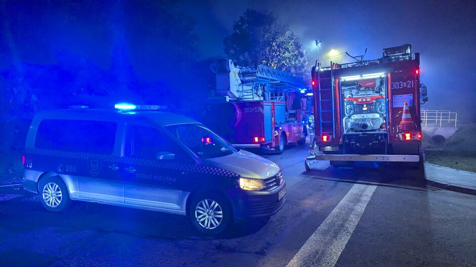 radiowóz straży miejskiej z włączonymi światłami niebieskimi, zaparkowany w poprzek ulicy, w tle dwa wozy gaśnicze straży pożarnej