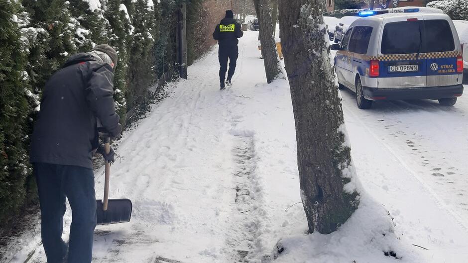 Mężczyzna odśnieża chodnik, w tle radiowóz i strażnik miejski.