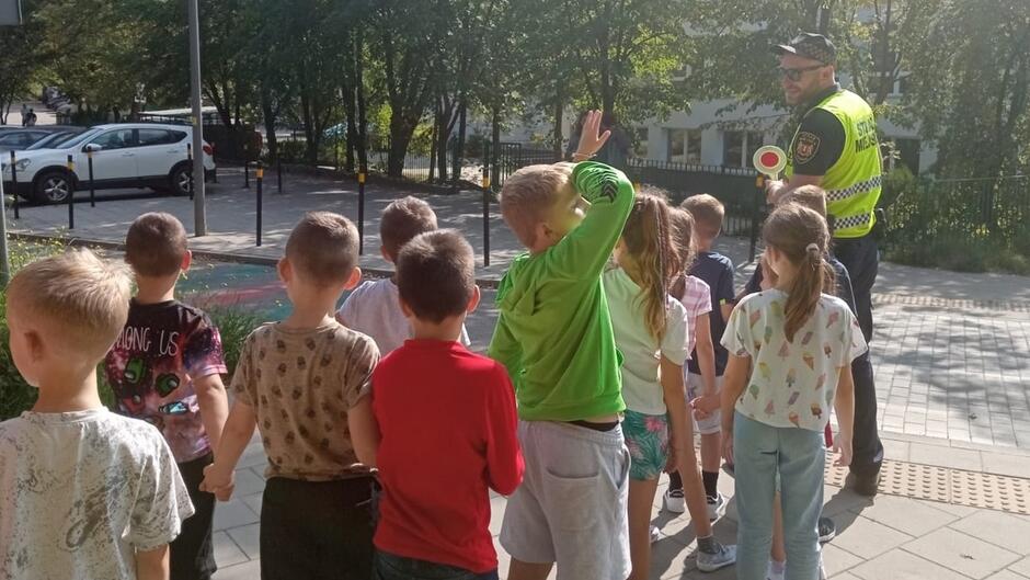 grupa dzieci i strażnik w żółtej kamizelce stoją na ulicy w tle samochody