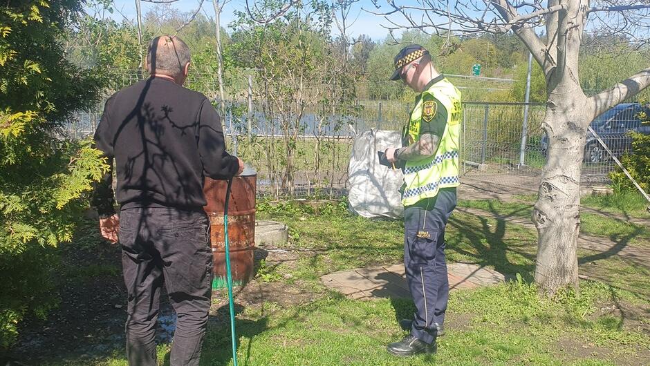 mężczyzna z wężem ogrodowym i strażnik stoją przy metalowej beczce na terenie ogródka działkowego w tle staw ogrodzenie i samochód