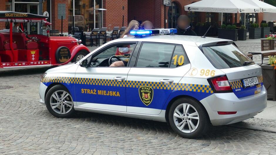 Na ulicy stoi radiowóz straży miejskiej, a przed nim meleks.