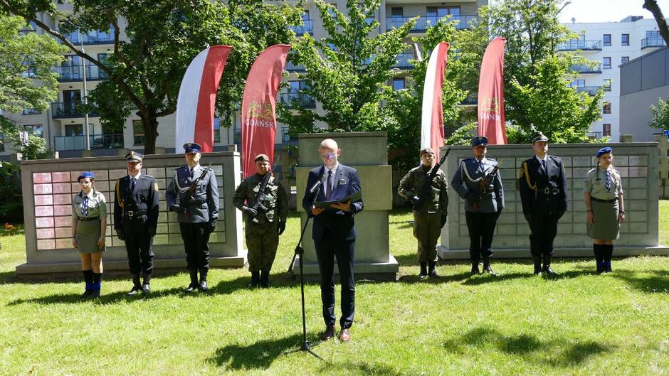 Przemawia Piotr Kryszewski, za nim pomnik i warta honorowa.