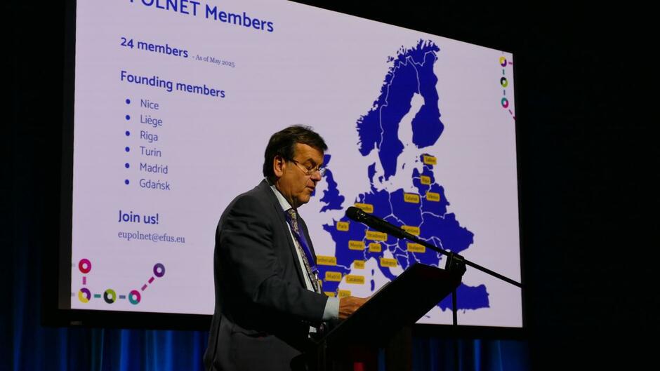 Przemawia Willy Demeyer, prezydent Efusu. Za nim plansza na temat sieci EU-POLNET.