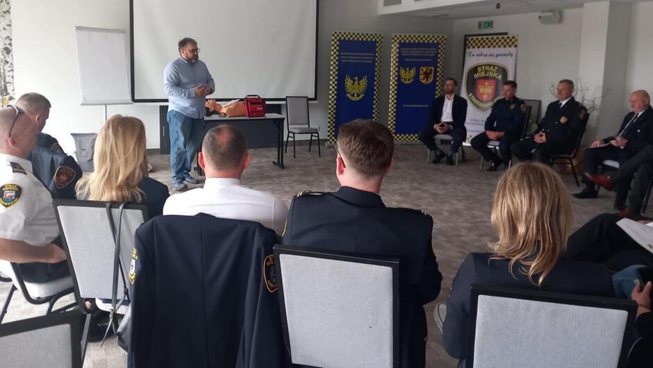 komendanci siedzą na krzesłach w tle banery instruktor pokazujący fantoma do pierwszej pomocy