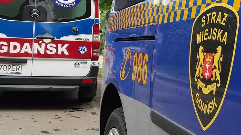 emblemat straży miejskiej w gdańsku na drzwiach radiowozu, w tle fragment zaparkowanej karetki