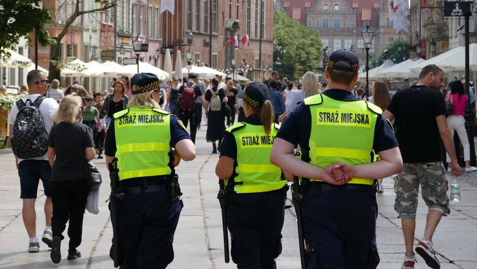 strażnicy idą ulicą Długą w kierunku Zielonej Bramy. wokół chodzą ludzie po bokach kamienice i drzewa