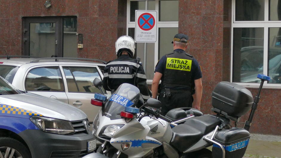 straznik miejski i policjant w kasku stoją plecami do aparatu. przy nich radiowoz straży i motocykl policjanta